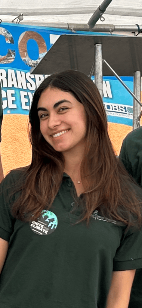 A young woman with long, dark hair smiles at the camera, wearing a dark green polo shirt and a button with an environmental theme, standing in front of a colorful backdrop.